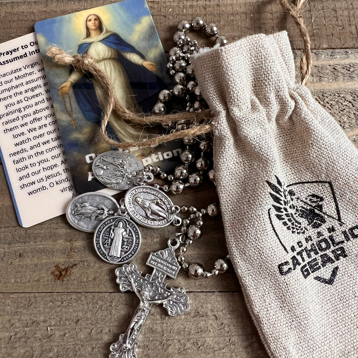 Silver Combat Rosary with an Our Lady of the Assumption prayer card and a Roman Catholic Gear drawstring bag on a wooden table
